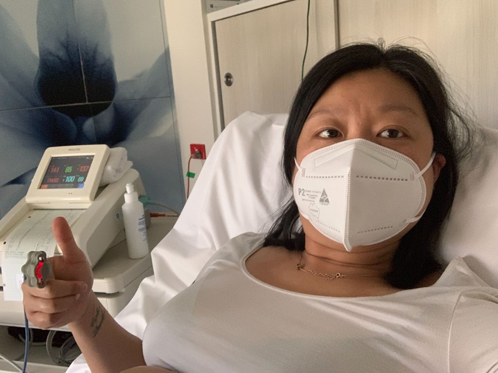 Natalie Khoo from Avion Agency smiling on a hospital bed during pregnancy with a COVID face mask on. 